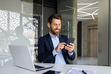 A successful and smiling young male businessman is sitting in the office at a desk in a suit and...