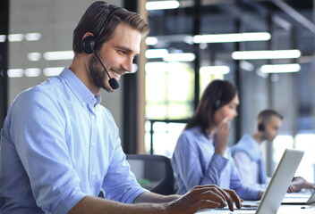 Smiling male call-center operator with headphones sitting at modern office with collegues on the backgroung, consulting online. - obrazy, fototapety, plakaty