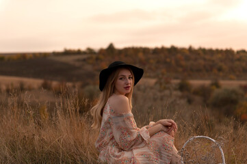 Young attractive woman in dress and hat sitting in grass on the hill at evening