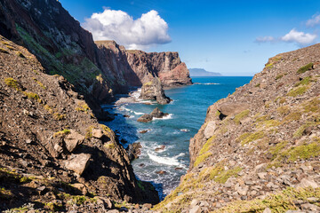 The photo depicts the rocky coast of Madeira. You can see the light blue sea, waves crashing against the rocks, and green vegetation on the slopes. It's a beautiful view of nature.