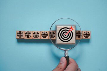 Hand holding magnifier glass to focus dartboard with arrow for business achievement objective goal and target concept.
