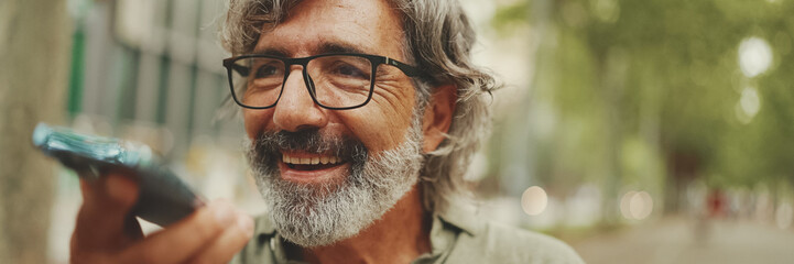 Clouse-up, Panorama of smiling middle-aged man with gray hair and beard sits on bench and uses mobile phone. Mature gentleman in eyeglasses recording voice recognition message on speakerphone - obrazy, fototapety, plakaty