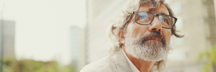 Clouse up portrait of mature businessman with beard in eyeglasses wearing gray pondering jacket...