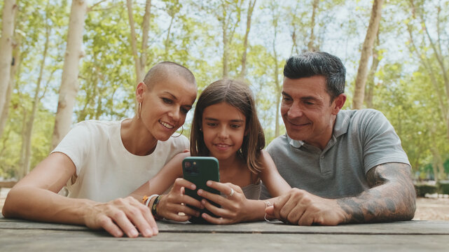 Happy family, mom dad and daughter, spending time together, family using smart phone looking at pictures and videos sitting in park ben