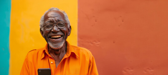 A man in an orange shirt is smiling and holding a cell phone. the man is enjoying his time with the phone. An elderly black man smiling and laughing with his phone against a colored background