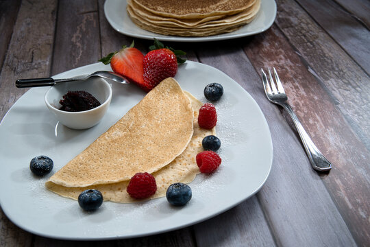 Pancakes with jam in a small bowl, blueberries, raspberries, strawberries sprinkled with powdered sugar. in the background are more pancakes on a plate.side view