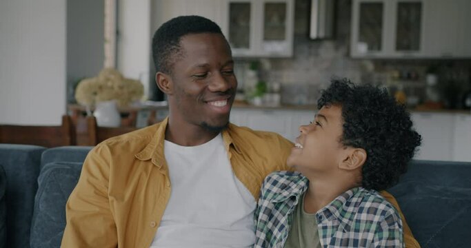 Joyful African American man and little boy son talking then looking at camera and smiling in apartment. Family relationship and happiness concept.