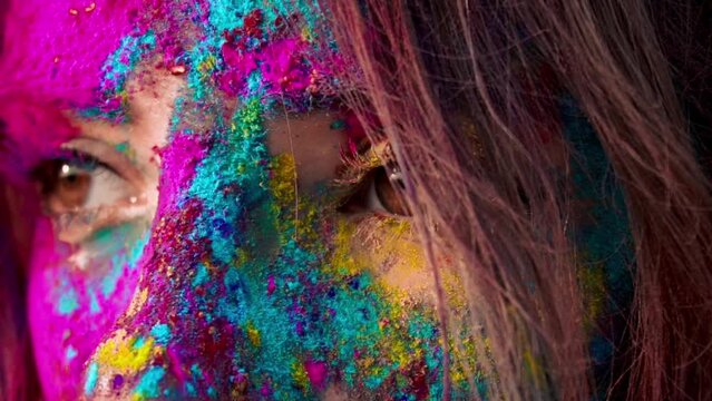 Female hazel brown eyes with colourful makeup open and close in slow motion closeup. Face covered with multi colorful powders. Celebrating Holi Festival. Creative abstract colorful splash art make-up.