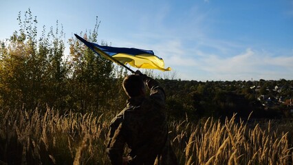 Male military in uniform waving flag of Ukraine at countryside. Young soldier of ukrainian army lifting blue-yellow banner as symbol of victory against russian aggression. Invasion resistance concept. - 766464600