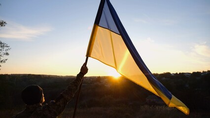 Male soldier stands with raised flag of Ukraine against background of beautiful sunset. Male ukrainian army soldier with a lifted blue-yellow banner in honor of the victory against russian aggression - 766464834