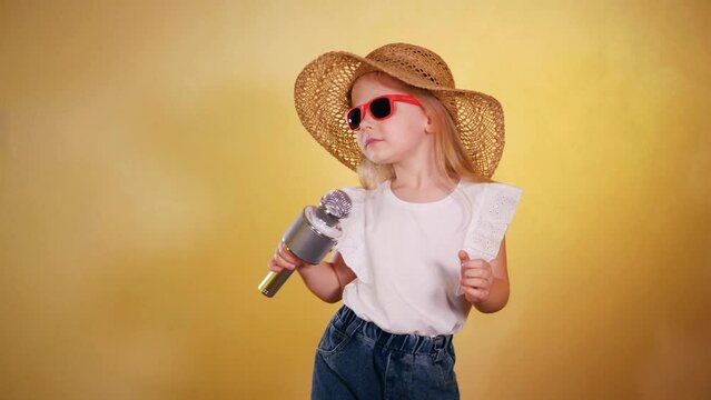 Little girl in hat and sunglasses singing into a microphone and dancing happily. Young girl in a fashionable hat and sunglasses belting out a song into a microphone. Child performer in stylish accesso