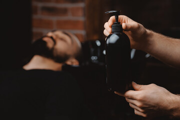 Mockup dark shampoo bottle on background of man in barbershop washing hair. Concept spa cosmetic...