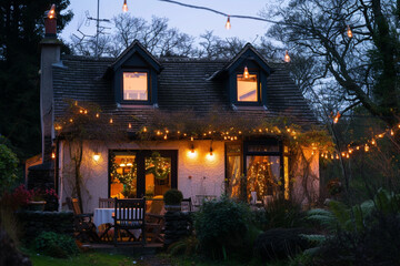 A charming cottage with fairy lights outside, set against a backdrop of mature trees, under the tranquil hues of a steel blue dusk