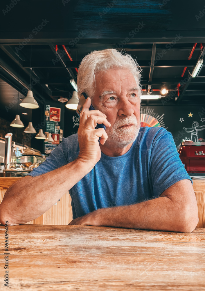 Wall mural Serious bearded senior white-haired man sitting at cafe table using mobile phone listening unpleasant news