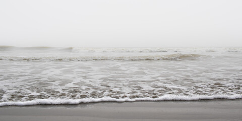 Waves of ocean on a Misty morning creating foam on sandy sea beach of grey color. 