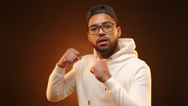 A stern-looking young man is showing a sign of aggression with his fists clenched, as if ready for a confrontation, his imposing stance set against a warm-toned background. Camera 8K RAW. 
