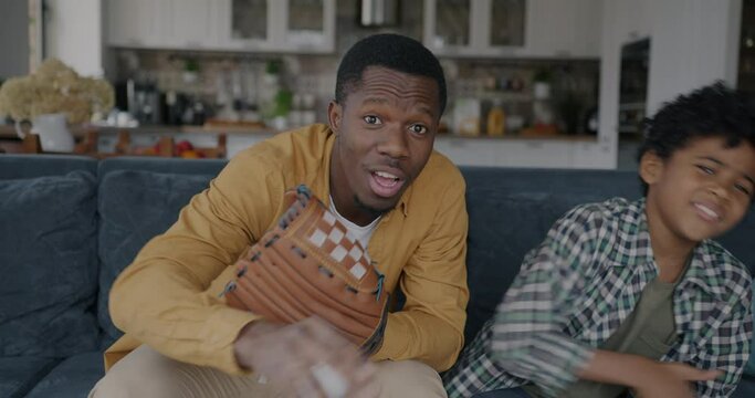 African American man watching baseball game with little son supporting cheering enjoying sports competition at home. Family and TV concept.