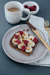 plate with toast with chocolate spread and banana, raspberry halves and nuts on a linen towel on a gray background. nearby are gold cutlery, a white cup of coffee and a bowl of raspberries
