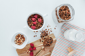 a golden spoon with granola on a wooden board, next to a bowl with raspberries and almonds. in the background there is a glass jar with granola and a carafe with milk and kitchen towel with stripes