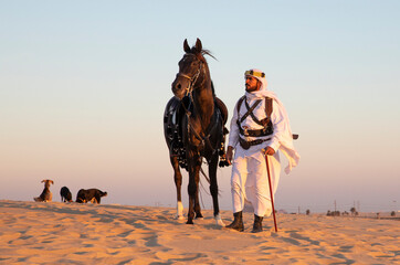 Saudi man  in traditional clothing in a desert at sunset with his horse