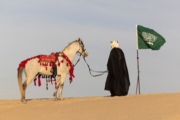 Saudi Man with his white stallion in a desert, carrying a flag of Saudi Arabia