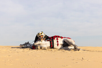 Saudi man in traditional clothing in a desert with his white stallion, aiming a hunting rifle