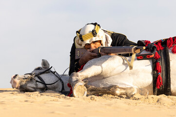 Saudi man in traditional clothing in a desert with his white stallion, aiming a hunting rifle