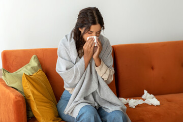 Sick young woman wrapped in blanket at home, blowing her nose with tissue