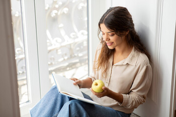 Beautiful young woman enjoying apple while reading interesting book at home - obrazy, fototapety, plakaty