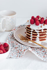 a stack of pancakes with vanilla sauce, decorated with raspberries and crushed almonds, behind a white cup with coffee and a bowl with raspberries. a colorful towel with gold cutlery. on a white backg