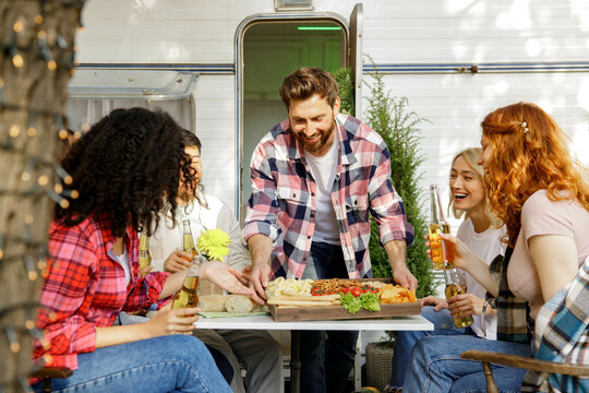 A cheerful multinational company is having fun on a picnic. Multisex friends sitting at the table outdoors celebrating eating and drinking drinks.