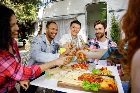 A cheerful multinational company is having fun on a picnic. Multisex friends sitting at the table outdoors celebrating eating and drinking drinks.