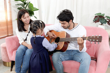 Happy Asian family. Chubby little girl daughter fell interested in acoustic guitar that dad...