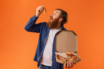 Redhaired bearded man in casual attire eats juicy pizza, studio