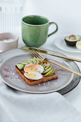 on two plates,toast with avocado and half an egg,next to golden cutlery.in the background there is a carafe with water,a green cup with coffee,a saucer with halves of avocado and eggs.white background