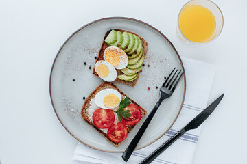 on a plate are two toasts with avocado and a boiled egg and with tomatoes and a boiled egg, next to them are black cutlery, a glass of orange juice and a kitchen towel. white background