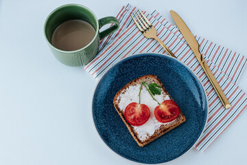 Fototapeta na wymiar on a blue plate there is toast with cream cheese and cherry tomatoes, next to it there is a striped kitchen towel and gold cutlery and a green cup of coffee. white background, shot from above