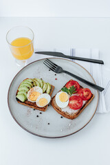 on a plate are two toasts with avocado and a boiled egg and with tomatoes and a boiled egg, next to them are black cutlery, a glass of orange juice and a kitchen towel. white background