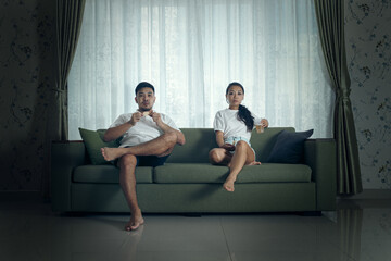A young couple dressed casually sits on a green couch against a curtained background, gazing pensively.