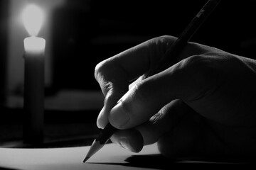 Close up of a man's hand writing on paper at night lit by candlelight, hand writing letter with pencil on paper in candle light condition. writing diary by candlelight in the dark. - obrazy, fototapety, plakaty