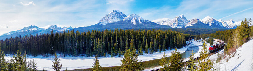 Panorama of Morant's Curve in Banff National Park as red cargo train passes through in Winter with Mount Temple and Paradise Valley in the background.