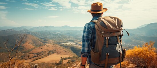 A traveler wearing a fedora and backpack stands atop a mountain under the sunny sky, admiring the tartan landscape and fluffy clouds - obrazy, fototapety, plakaty