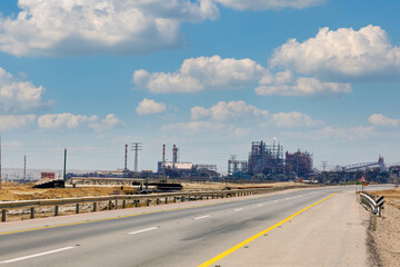 Open highway leading towards an industrial area. The road is empty and marked with yellow lines. Plant for the production of magnesia from Dead Sea salts with smoking chimney. Ein Tamar, Israel.