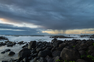Fototapeta na wymiar Long exposure shot of the waves crashing on the rocky shore and the rain clouds approaching the coast of Benares beach in a morning on the south coast of Mauritius island