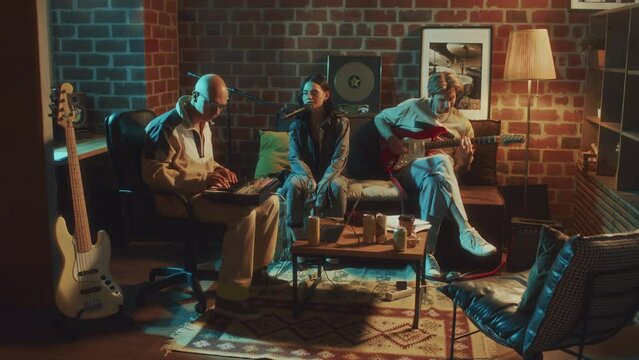 Full footage of Caucasian indie musical band creating or rehearsing rock song to electric guitar and using beat maker machine in vintage-style living room with soft light in evening