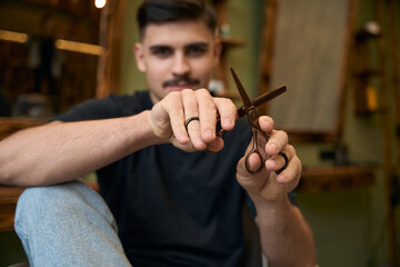 Barber showing scissors while sitting at his workplace in barbershop