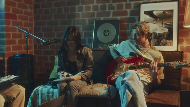 Panning footage of Caucasian female and male musicians creating music on guitar and midi keyboard while sitting in retro studio in evening