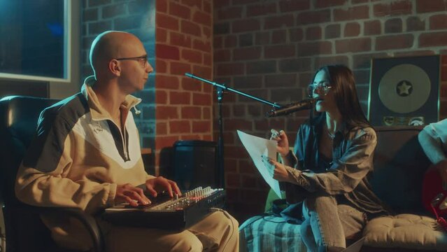 Panning softlight footage of Caucasian friends creating music together in retro studio, men playing guitar and beat makeing while lady signing lyrics in evening