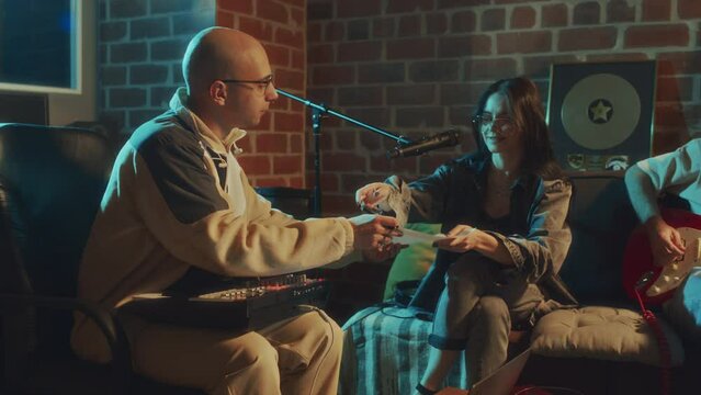 Side footage of Caucasian male beat maker passing paper with lyrics to female vocalist who checking mic and then discussing song production while rehearsing in retro studio in evening