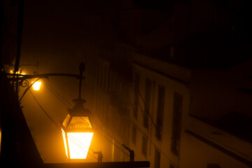 Mysterious Foggy Night in Old Town with Vintage Street Lamp. A warm glow emanates from an antique...